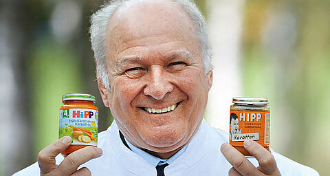 An elderly man with a white shirt holds two jars of baby food, one with a label that reads "HIPP Karotten" and the other with "HIPP Frukt-Karotten-Gemüse" and a depiction of a baby.