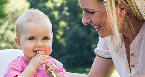 A woman and a baby are outdoors, with the woman looking at the baby who is chewing on a spoon.