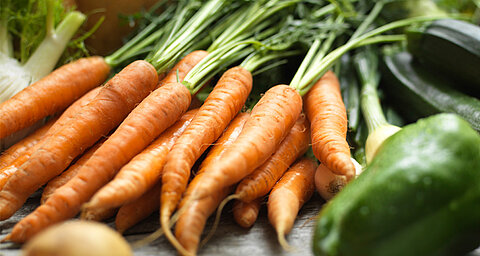 A close-up of fresh, vibrant orange carrots with green tops, surrounded by other vegetables.
