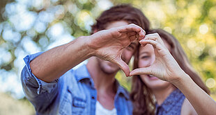 Two people, one male and one female, are holding their hands together in a heart shape, with the female's face partially visible.