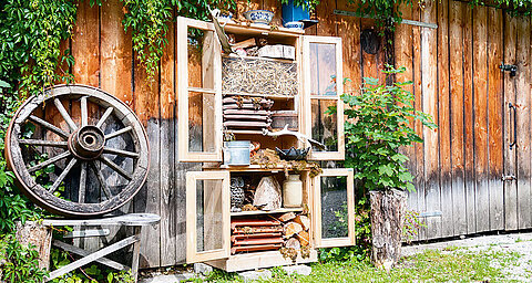 A rustic wooden shelf stands against a weathered wooden wall, filled with various items, next to a vintage wagon wheel.