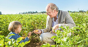 An elderly man and a young child are in a field, with the man holding a potato and handing it to the child.