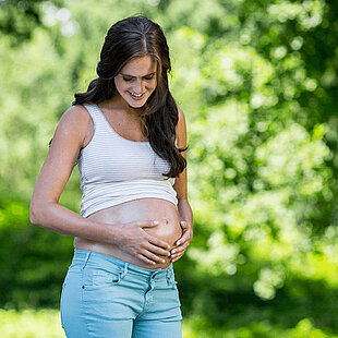 A pregnant woman in a white and gray striped tank top and blue jeans stands outdoors, looking down at her belly.