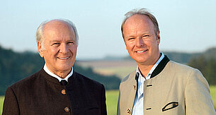 Two men, one older with white hair and the other younger with brown hair, stand side by side outdoors.
