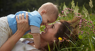 A woman and a baby are lying on grass, surrounded by flowers, with the woman holding the baby close to her face.