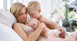A woman is lying on a bed, holding a baby and a toddler.