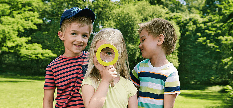 Three children play in a park; one peers through a magnifying glass.
