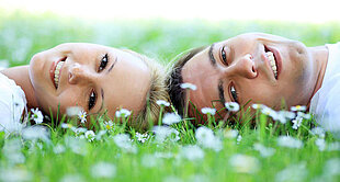 Two individuals lie on a grassy field with white flowers, looking up towards the camera.