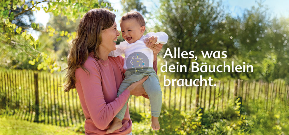 A woman in a pink shirt holds a laughing baby in a white shirt and green pants, with a fence and greenery in the background.