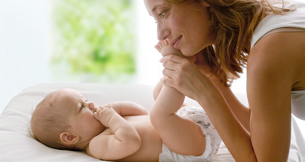 A woman in a white tank top is lying on a white bed, holding a baby's foot, with a blurred green background.