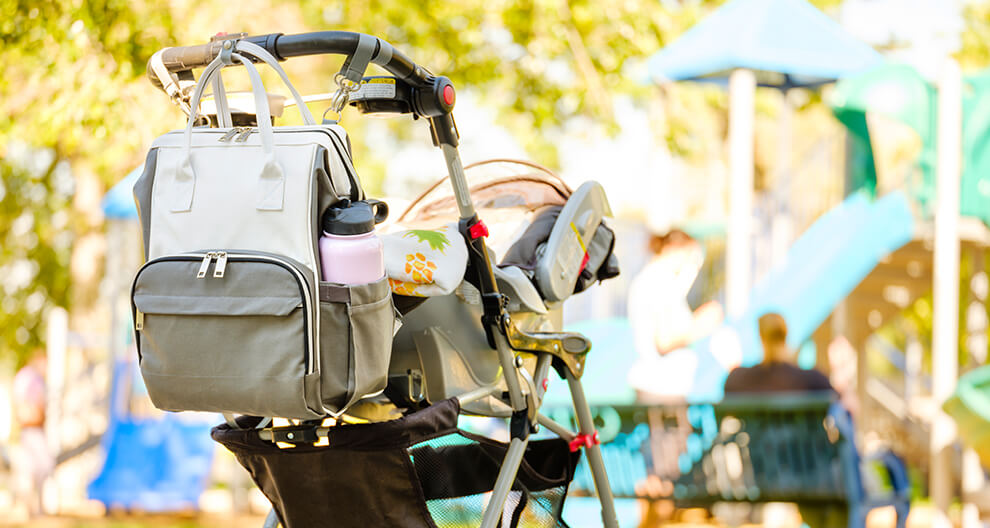Stroller with attached bag and water bottle, in a park with playground equipment and people in the background.