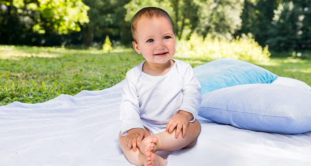 A baby sits on a white blanket outdoors.