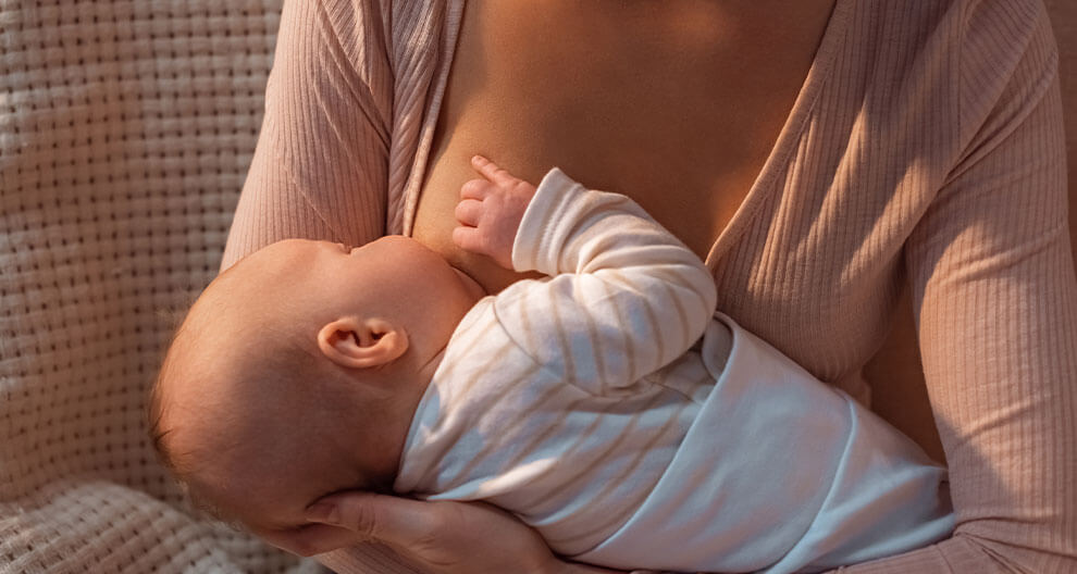 A woman in a beige top is breastfeeding a baby dressed in white and beige striped clothing.
