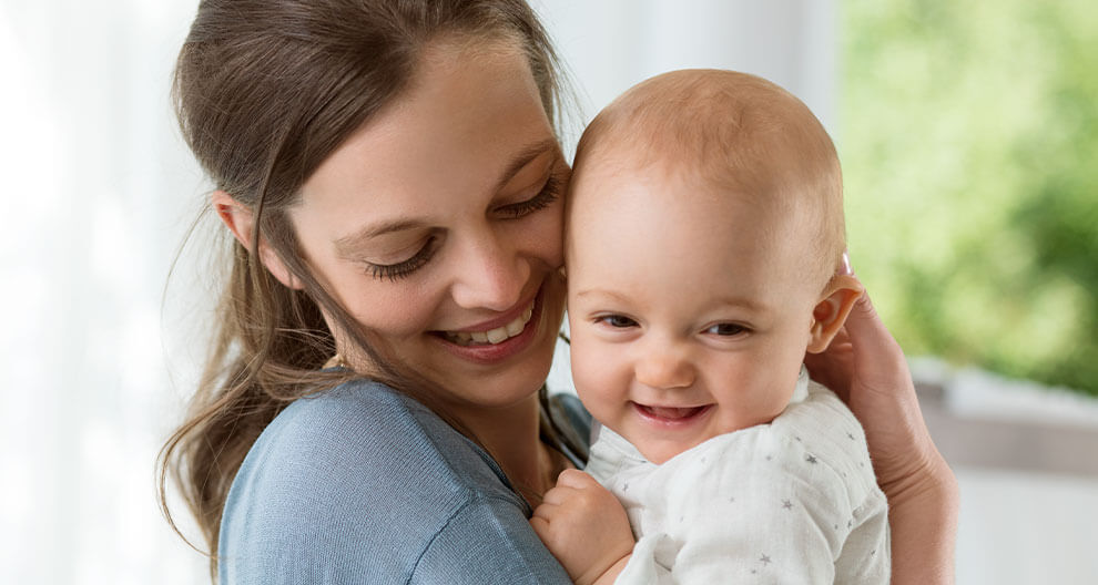 A woman in a blue shirt holds a baby with a white outfit, both looking at the camera.