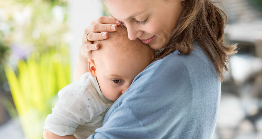 A woman in a blue shirt is holding a baby close to her, with the woman's face close to the baby's head.