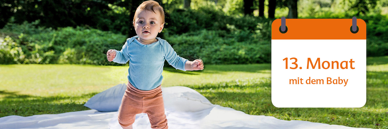 A baby in a blue shirt and brown pants stands on a white sheet in a grassy area with trees in the background, next to a calendar that reads "13. Monat mit dem Baby" in German.