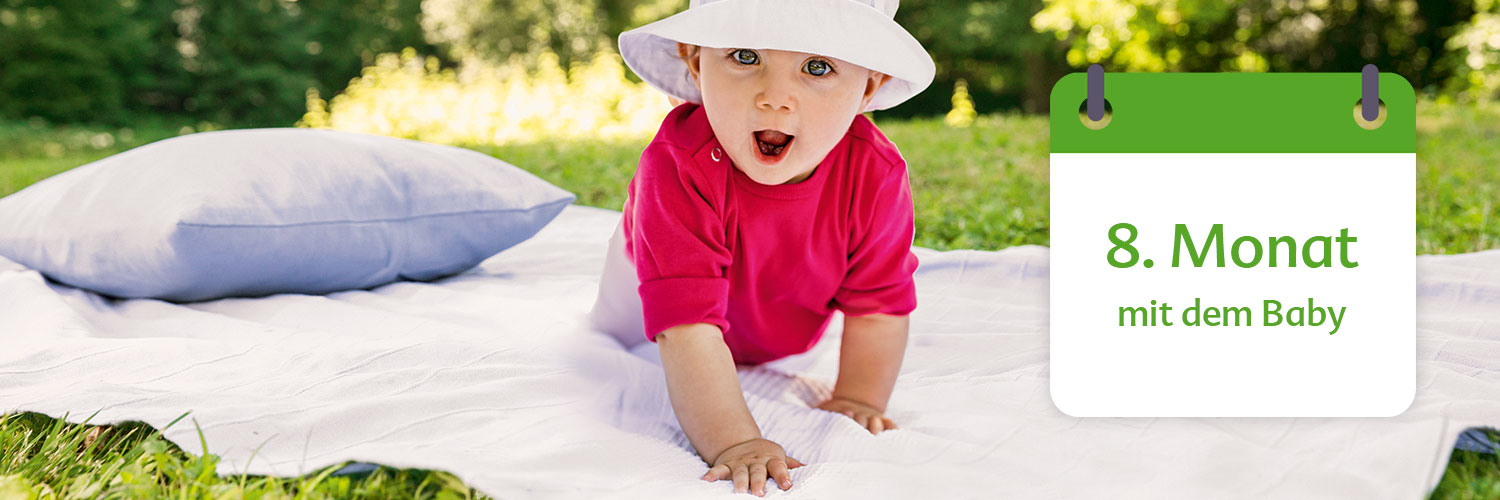 A baby in a red shirt and white hat is crawling on a white blanket in a grassy area with a pillow nearby, accompanied by a green calendar indicating the 8th month with the text "mit dem Baby" (with the baby).