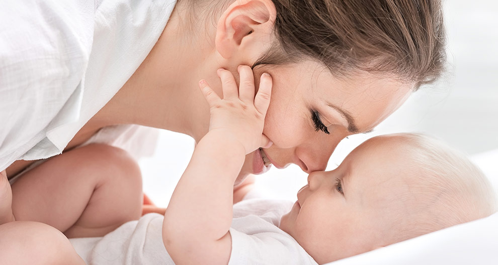 A woman in white clothing is lying down, cradling a baby who is reaching out to touch her face.
