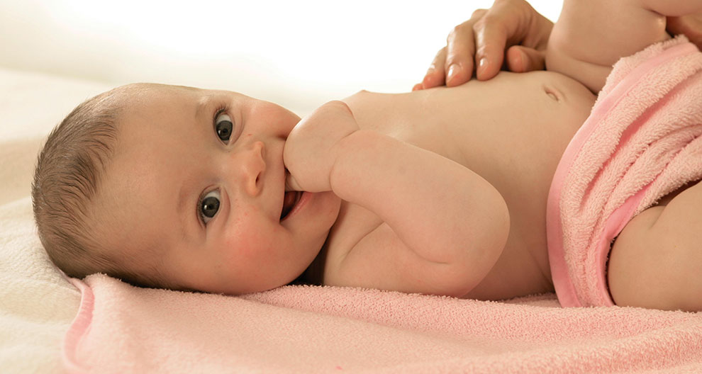 A baby lies on a pink towel, partially covered by a pink cloth, with a hand placed on the baby's leg.