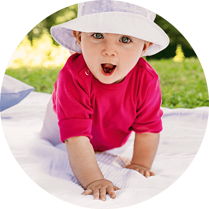 A baby wearing a red shirt and a white hat is crawling on a white blanket in a grassy area.