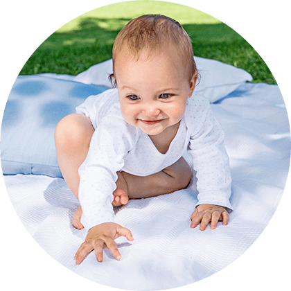 A baby in white clothing is crawling on a white blanket outdoors.