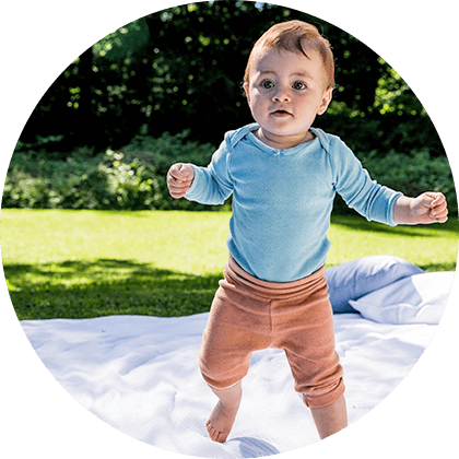 A baby in a blue shirt and brown pants stands on a white sheet in a grassy area with trees in the background.