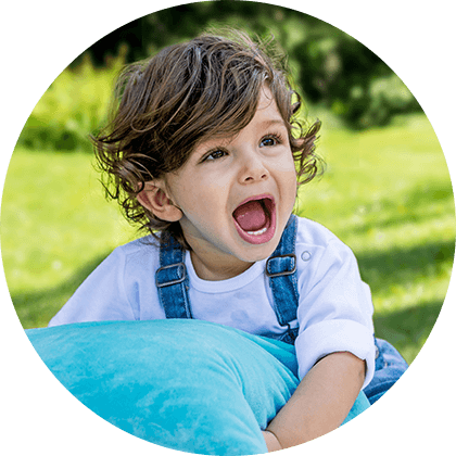 A child with curly hair, wearing a white shirt and blue overalls, is holding a blue pillow and appears to be shouting or expressing surprise.
