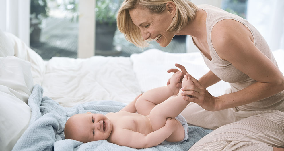A woman in beige attire is seated on a white bed, holding a laughing baby's feet.