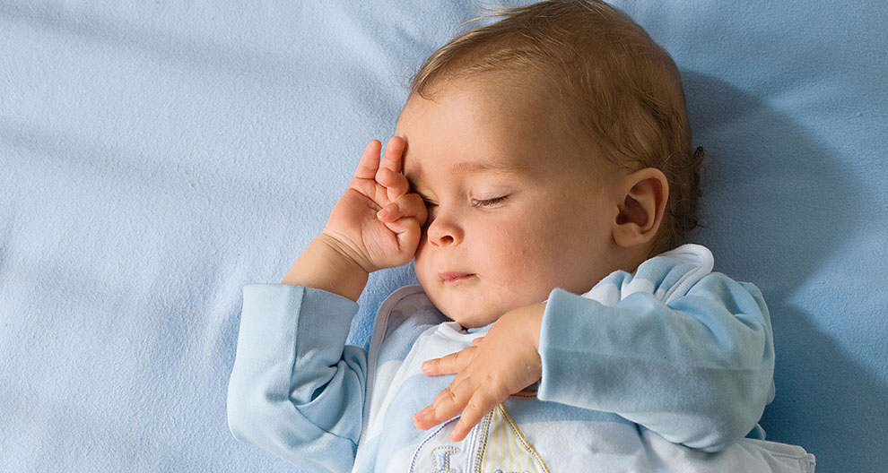 A baby lies on a blue surface, wearing a light blue outfit with a sailboat design, and covering one eye with a hand.