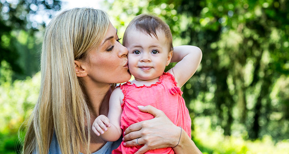 A woman with long blonde hair is kissing a baby wearing a pink dress.