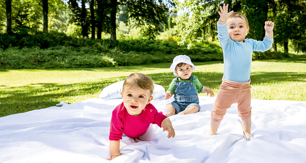 Three toddlers are playing on a white blanket in a grassy park.