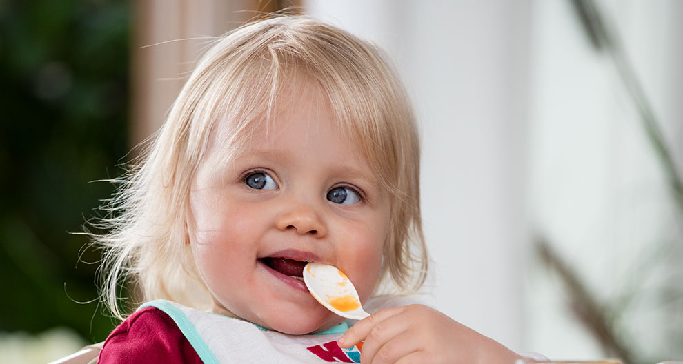 A young child with blonde hair is eating with a spoon.