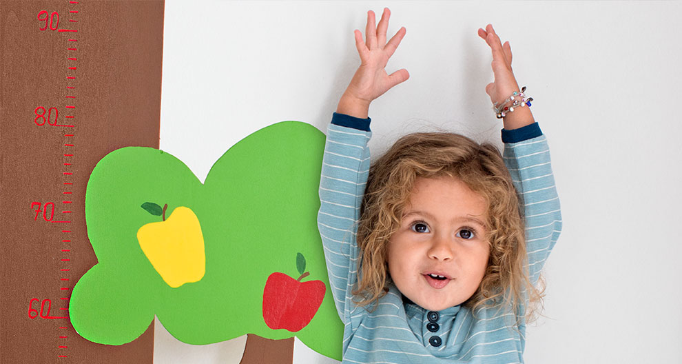 A child stands against a height measurement chart, arms raised, with a tree illustration and fruit depicted on the chart.