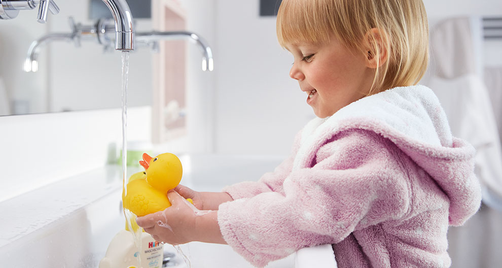 A child in a pink robe is washing a yellow rubber duck under a silver faucet.