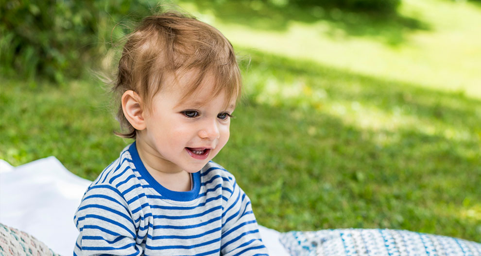 A toddler in a striped shirt sits outdoors on a blanket.