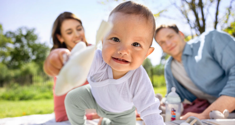A baby reaching out with a toy, with a man and woman in the background.