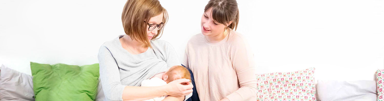 Two women and a baby are in a room with a green pillow and a floral pillow.