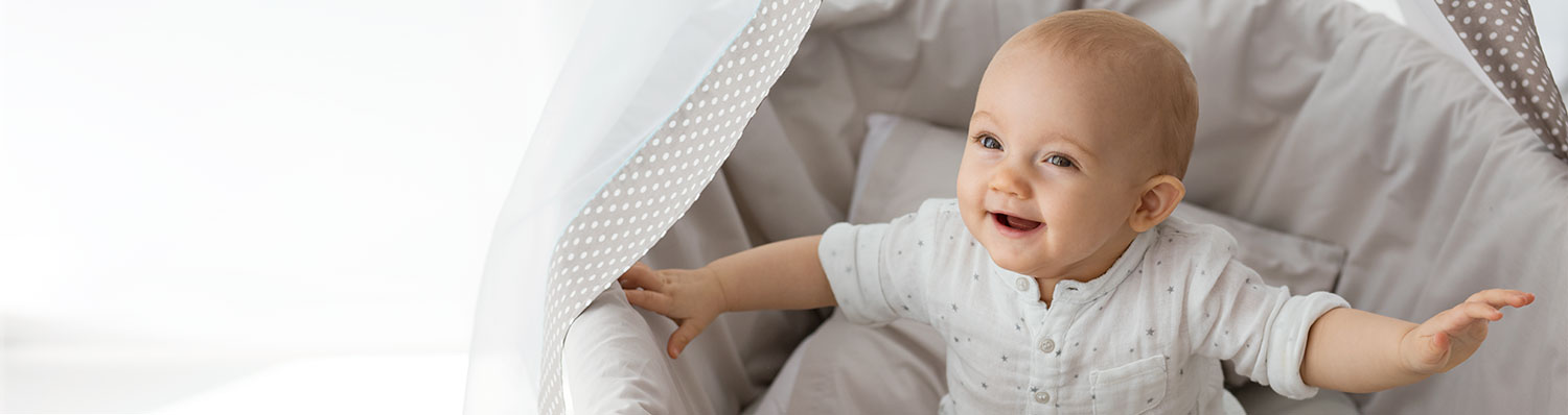 A baby in a white outfit is lying in a crib with a gray and white polka-dotted sheet.