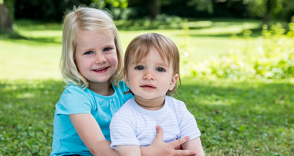 Two children, a girl in a blue shirt and a boy in a striped shirt, are sitting on grass in a park.