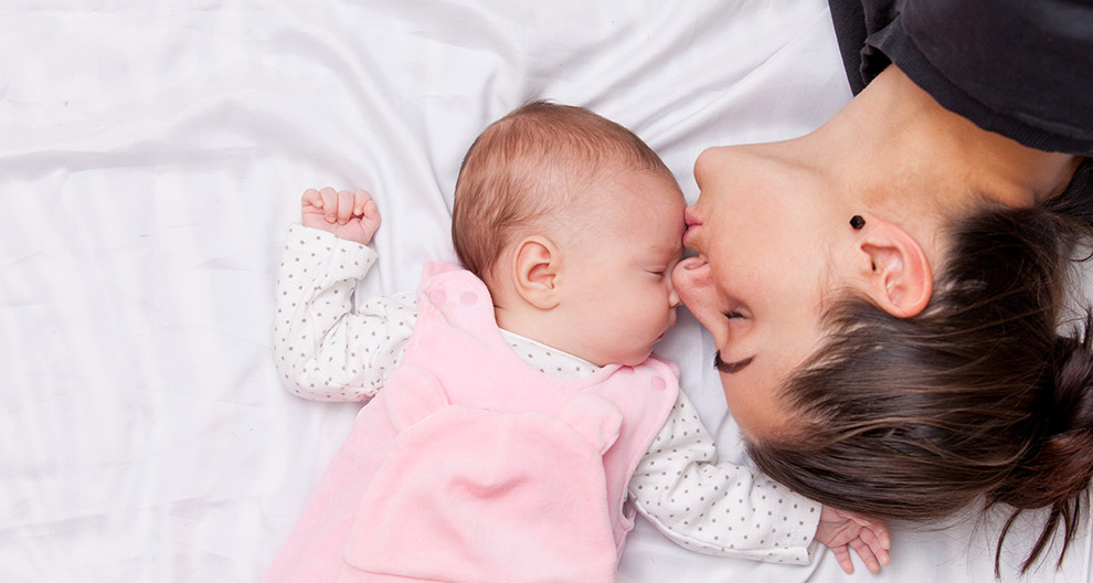 A woman and a baby are lying on a white bed, with the woman leaning over to kiss the baby's forehead.