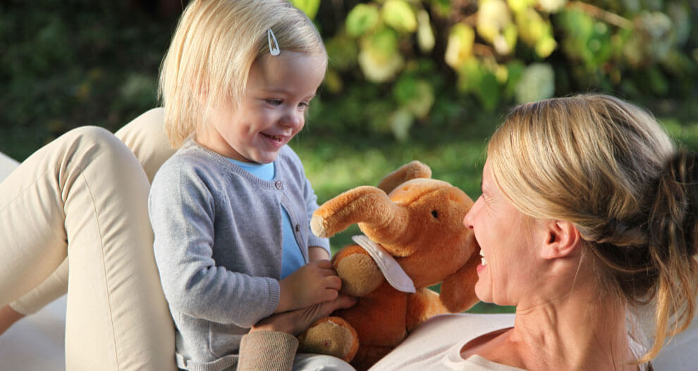 A woman and a young girl share a moment of joy and connection outdoors, with the woman holding a plush elephant toy.