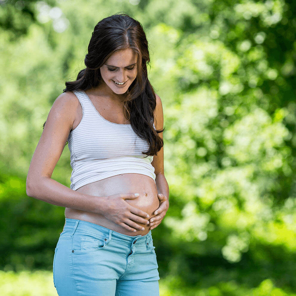 A pregnant woman in a white and gray striped tank top and blue jeans stands outdoors, looking down at her belly.