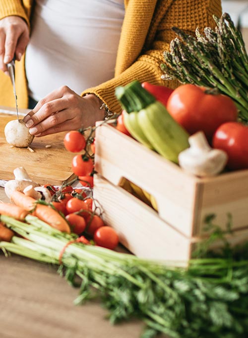 A person is cutting a mushroom on a wooden cutting board with a variety of fresh vegetables around.