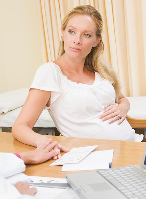 A woman in a white shirt sits at a table, holding a document, with a laptop in front of her.