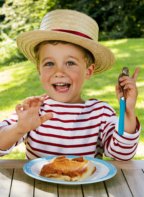 A child in a striped shirt and straw hat is excitedly holding a spoon, with a dessert on a plate in front of them.