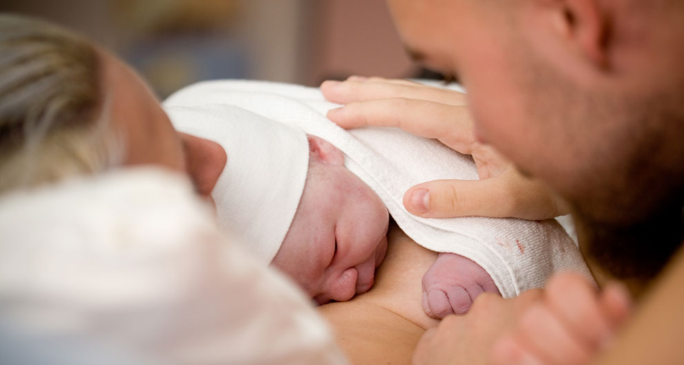A newborn baby is cradled by two adults, with one adult's face partially visible.