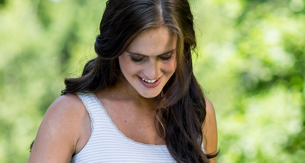 A woman with long brown hair, wearing a white tank top, is smiling and looking down.