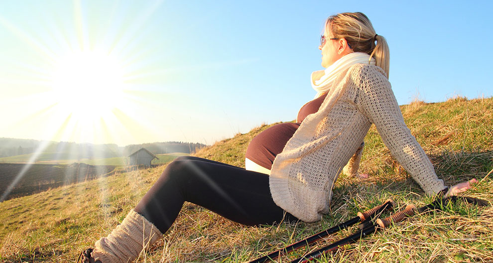 A pregnant woman sits on a grassy hill, looking at the sun, with walking poles beside her.