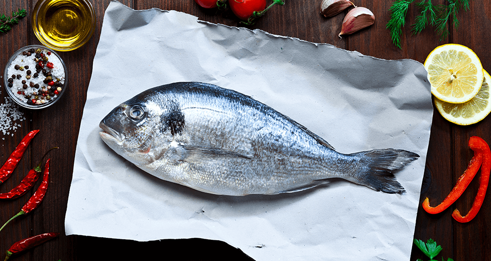 A fresh fish rests on a parchment paper surrounded by vibrant ingredients.