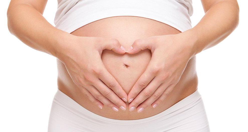 A pregnant woman forms a heart shape with her hands over her belly.